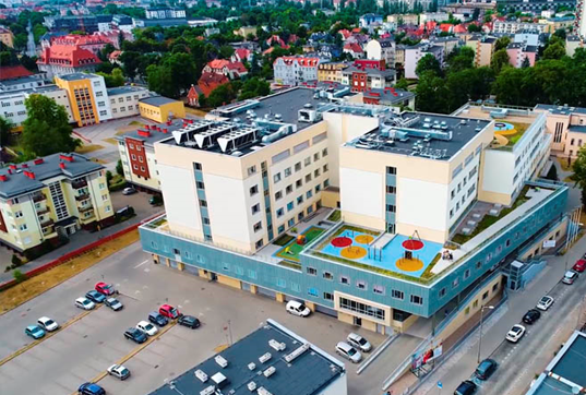 Budynek szpitala z lotu ptaka z widocznym parkingiem przed szpitalem i placami zabaw na dachu budynku