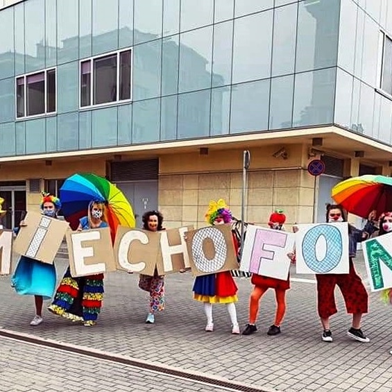 Wolontariusze z Fundacji Dr Clown prezentują kolorowy napis „Śmiechofon” na tle budynku szpitala