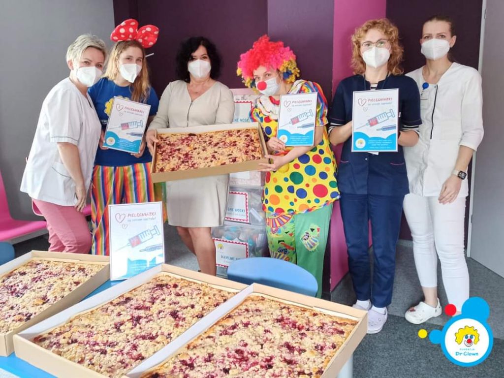 Zdjęcie przedstawia pracowników służby zdrowia i clowna, którzy celebrują dzień pielęgniarek, trzymając torty owocowe.