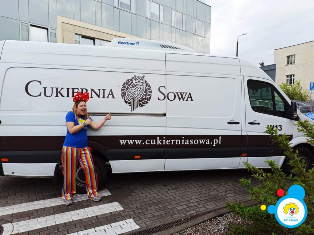 Kobieta w kolorowym stroju robi kciuka w górę obok furgonetki Cukierni Sowa, oferującej smakołyki.