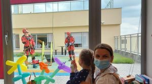 Dzieci i dorosły w maseczkach, obserwują ratowników w odzieży ochronnej na zewnątrz. Kolorowe balony w tle.