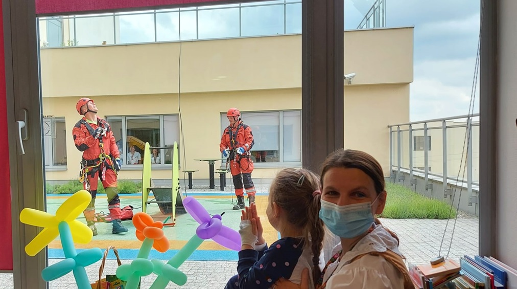 Dzieci i dorosły w maseczkach, obserwują ratowników w odzieży ochronnej na zewnątrz. Kolorowe balony w tle.