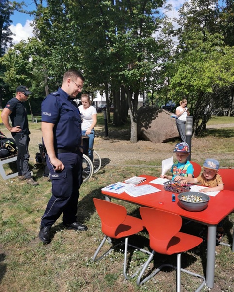 Policjant rozmawia z dziećmi przy stole w parku, podczas wydarzenia społecznego z rodzinami.
