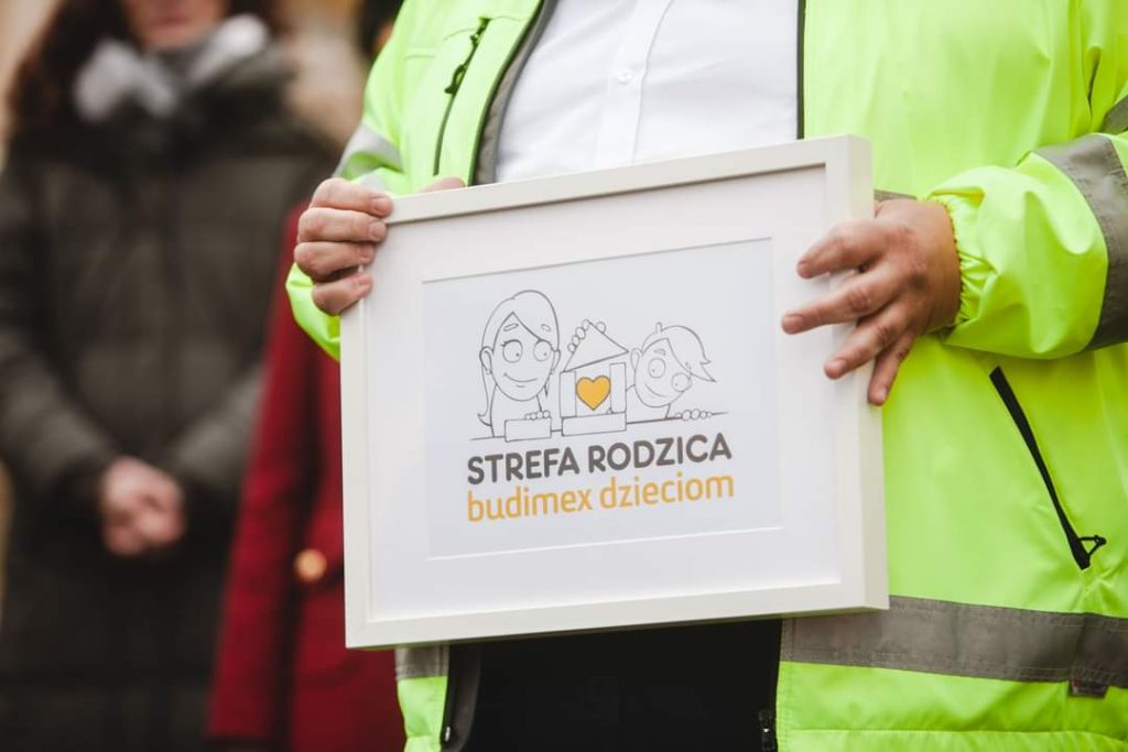 Osoba w żółtej kamizelce trzyma ramkę z logo "Strefa Rodzica - budimex dzieciom", promującym projekt dla dzieci.