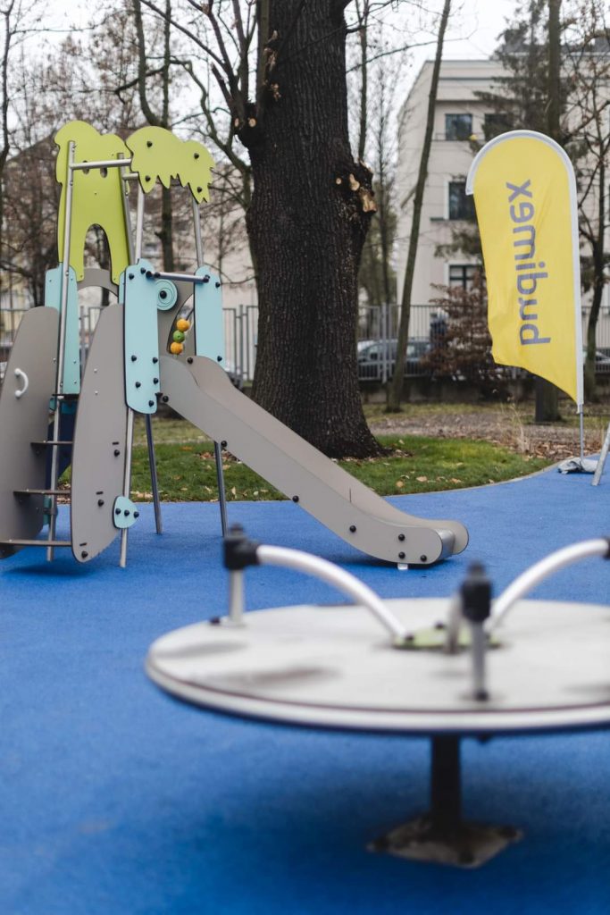 Nowoczesny plac zabaw z plastikową zjeżdżalnią i karuzelą, otoczony zielenią. 