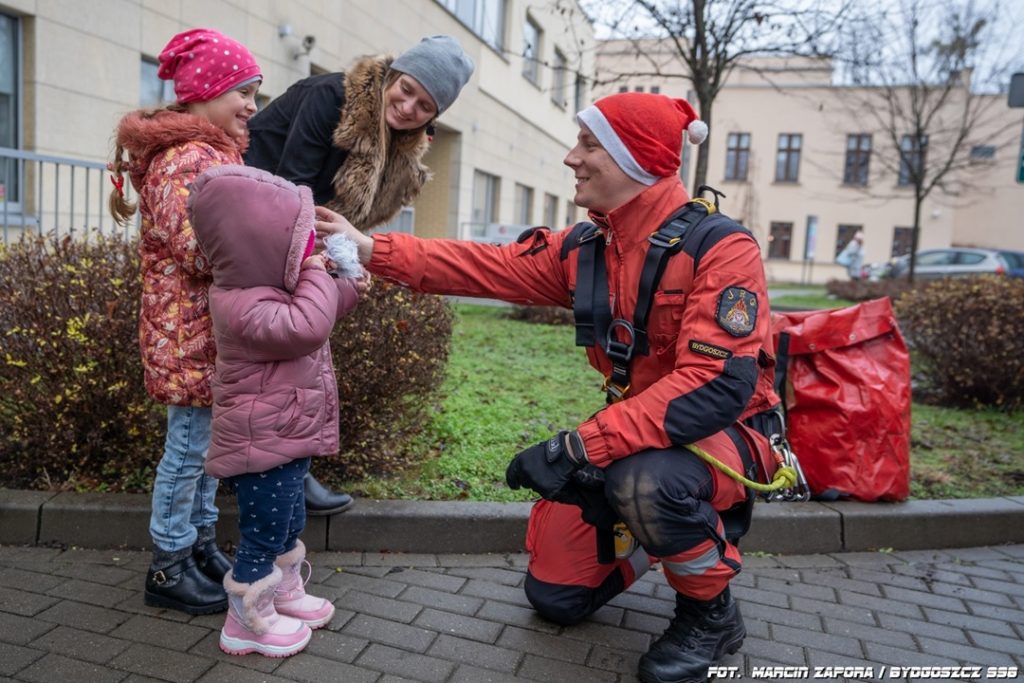 Strażak w czapce św. Mikołaja rozmawia z dziećmi przed budynkiem szpitala