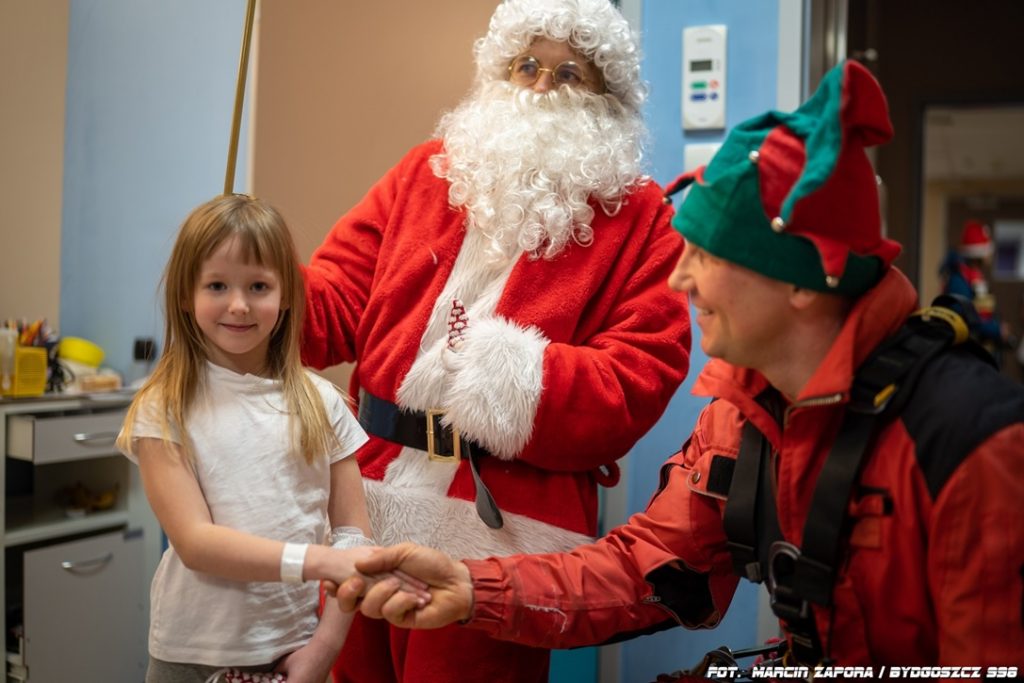 Dwójka strażaków – jeden w stroju św. Mikołaja, drugi w stroju Elfa witają się z pacjentką w
oddziale szpitalnym