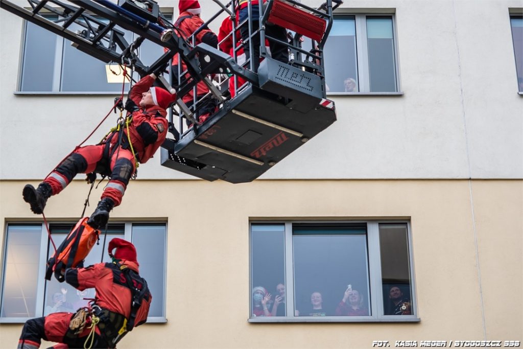 Pacjenci w oknach szpitala pozdrawiają strażaków wspinających się na gmach budynku
szpitala