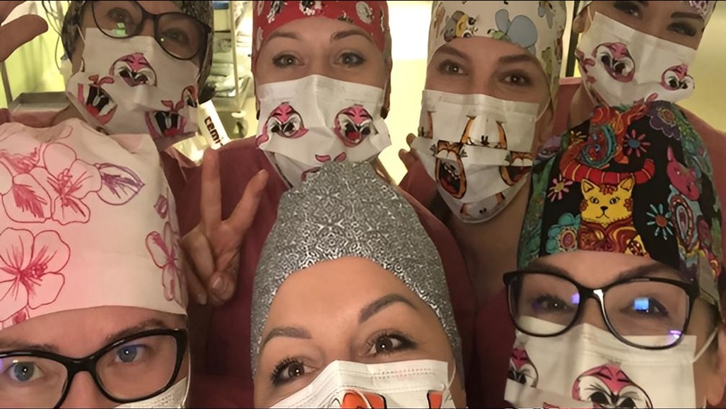 Grupa uśmiechniętych pracowników służby zdrowia w kolorowych czepkach i maseczkach ochronnych, wykonujących selfie w szpitalu.