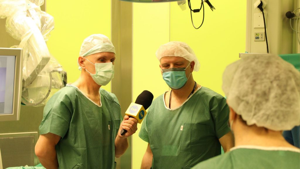 Dziennikarz przeprowadza wywiad z lekarzami w sali operacyjnej, wszyscy w zielonych fartuchach i maskach.