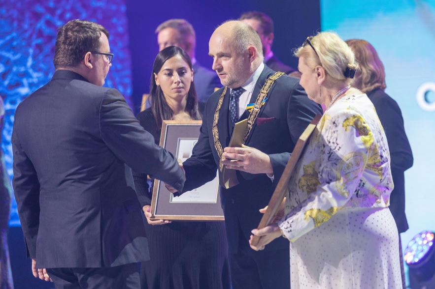 Uczestnicy ceremonii wręczenia nagród, mężczyzna w garniturze wita się z laureatem.