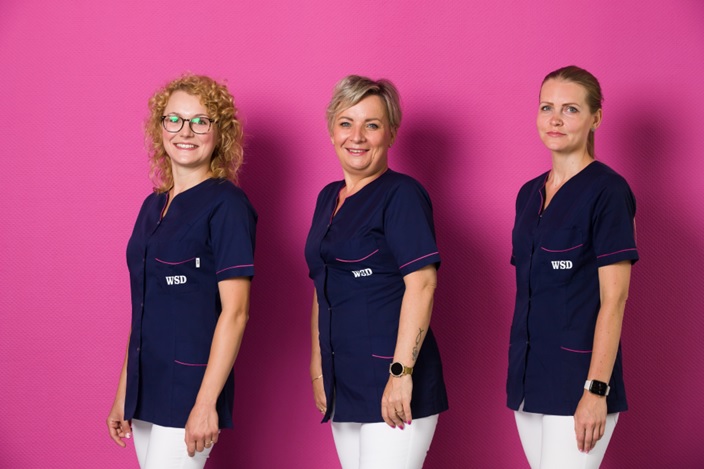 Trzy uśmiechnięte pracownice dietetyki w mundurach WSD na tle różowej ściany.