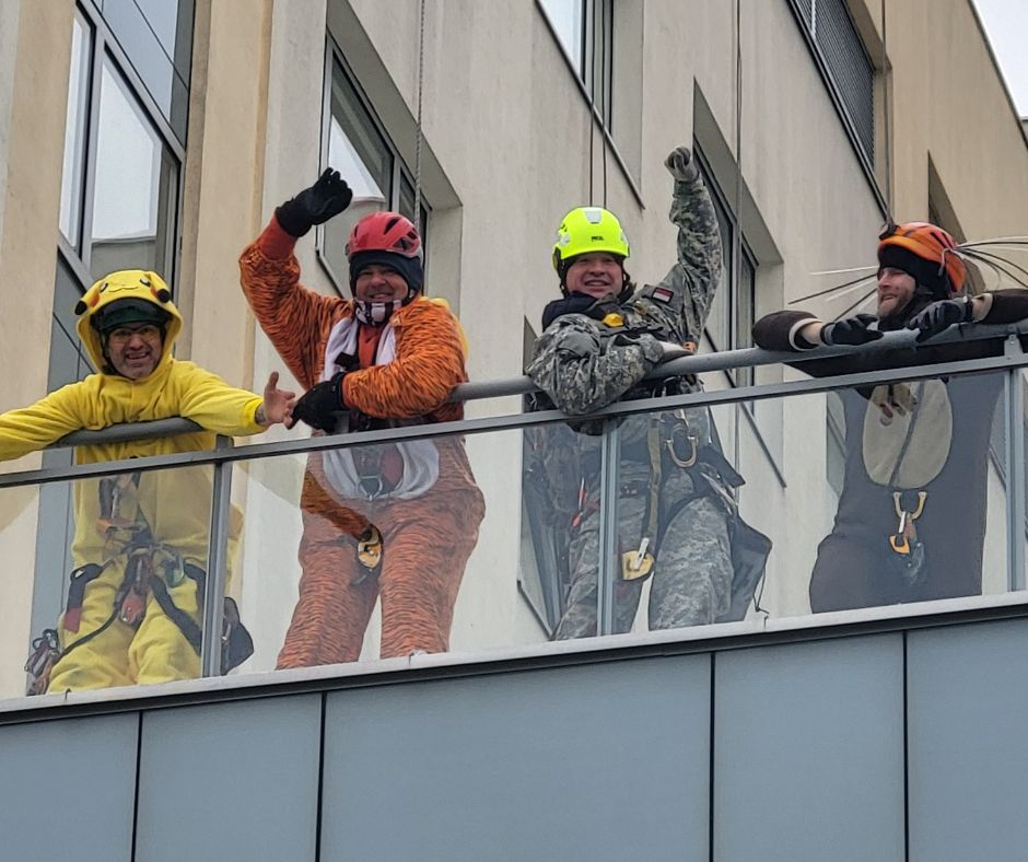 Alpiniści na dachu szpitala machają do obiektywu: od lewej alpinista w żółtym stroju pokemona Pikachu, alpinista w pomarańczowym stroju tygrysa, alpinista w wojskowym stroju moro, alpinista w brązowym stroju niedźwiedzia 