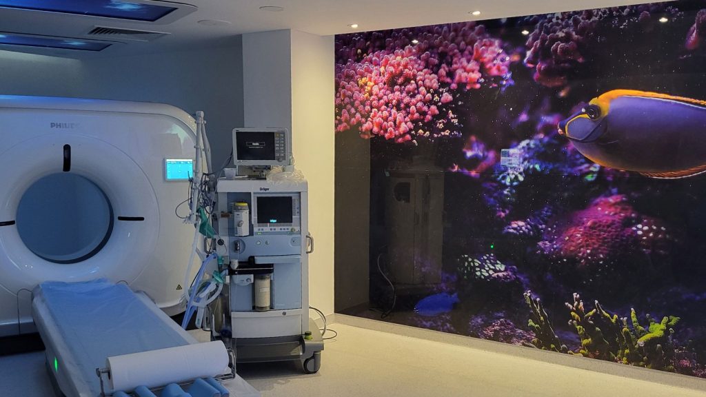 Aparat Incisive CT Philips Heathcare w Pracowni Tomografii Komputerowej, obok aparatu podświetlana ściana – zdjęcie rafy koralowej i ryb, na suficie projekcje kolorowych animacji, które pacjent ogląda w trakcie badania