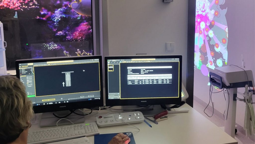 Technik obserwuje na monitorach komputerów przebieg badania, za szybą widać pomieszczenie z tomografem