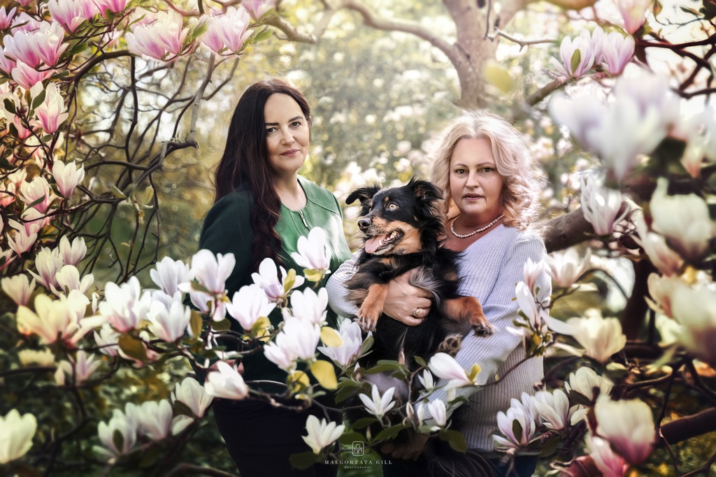 Dwie kobiety trzymające psa, w otoczeniu różowych magnolii, w scenerii natury.