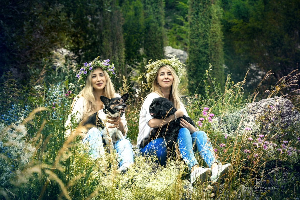 Dwie uśmiechnięte kobiety w wiankach, siedzące wśród kwiatów z psami na rękach.