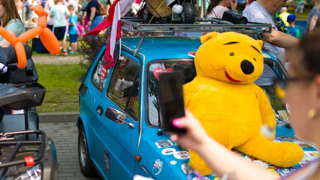 Niebieski mały fiat z pluszowym Kubusiem Puchatkiem na masce, w tle uczestnicy festynu